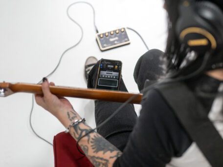 Positive Grid SPARK PEDAL Positive Grid SPARK PEDAL liegt auf dem Boden, Frau spielt und Smarphone mit Spark App auf dem Bein liegen hat.